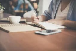 Persona escribiendo en un cuaderno junto a una taza de café y un móvil