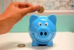 Mano echando una moneda en una hucha azul con forma de cerdito